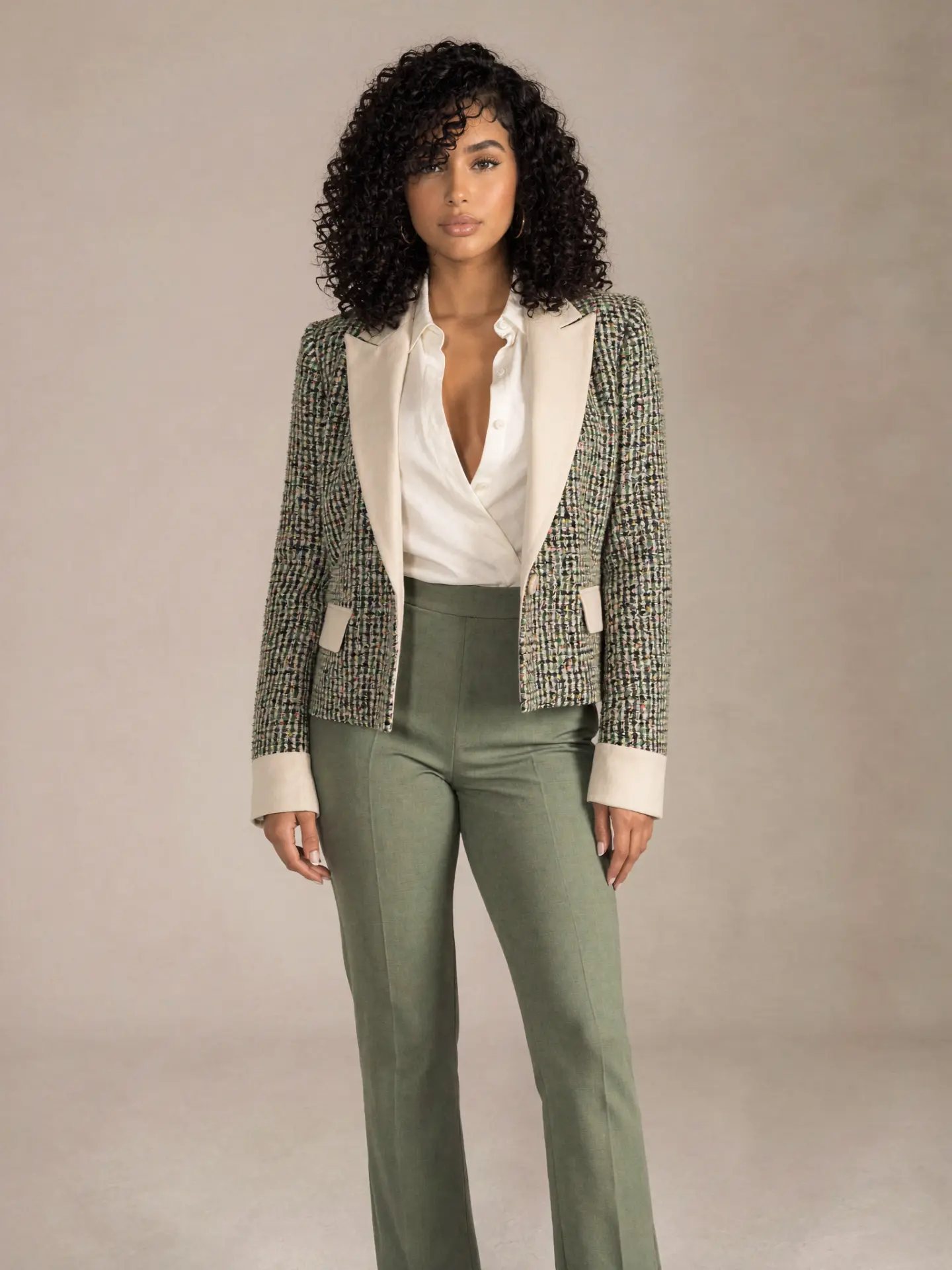 Portrait of Nocturna, a woman with long curly hair wearing a textured tweed jacket and olive green trousers against a warm neutral backdrop